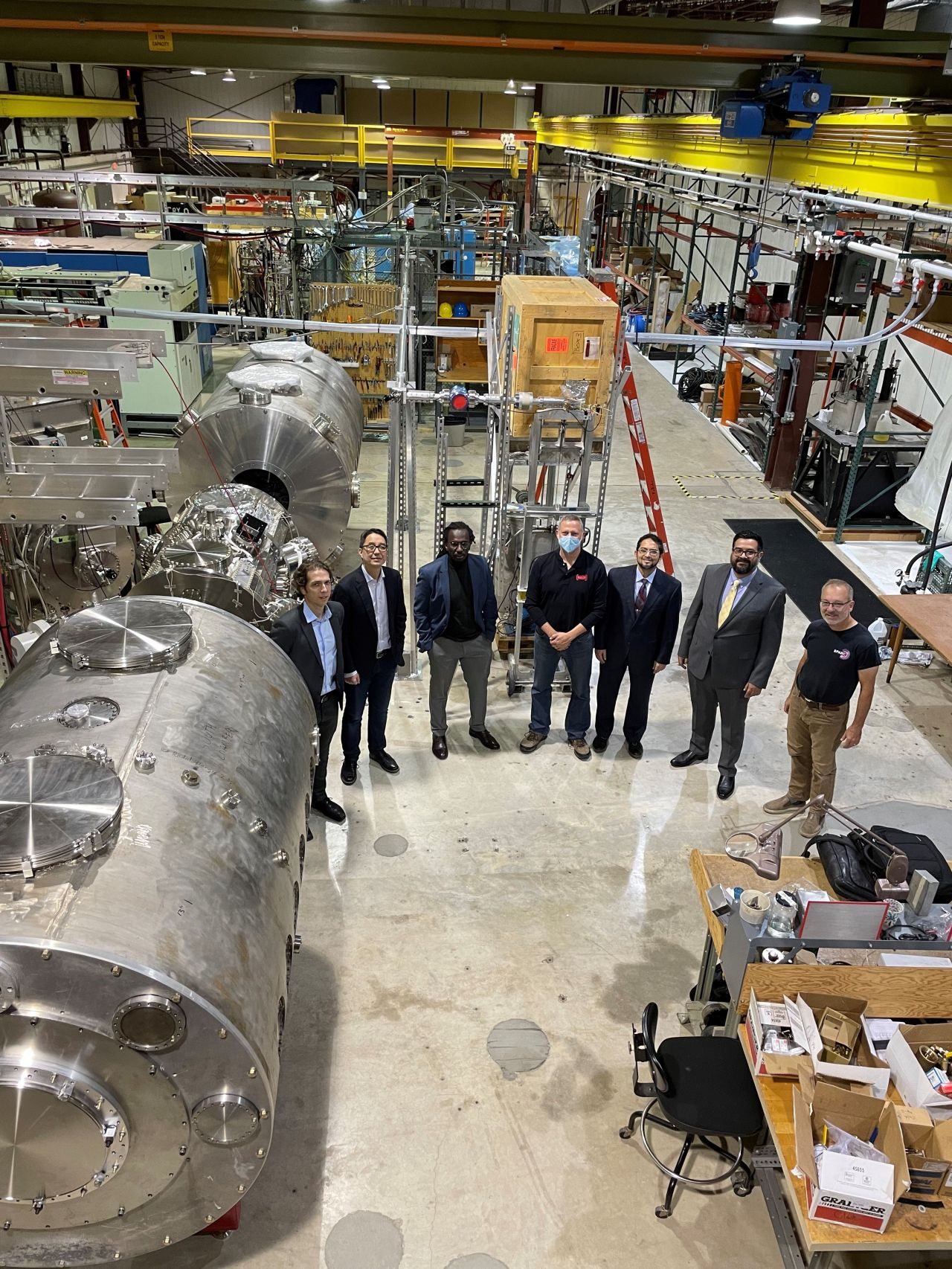 seven men stand in front of a large piece of scientific equipment in a warehouse