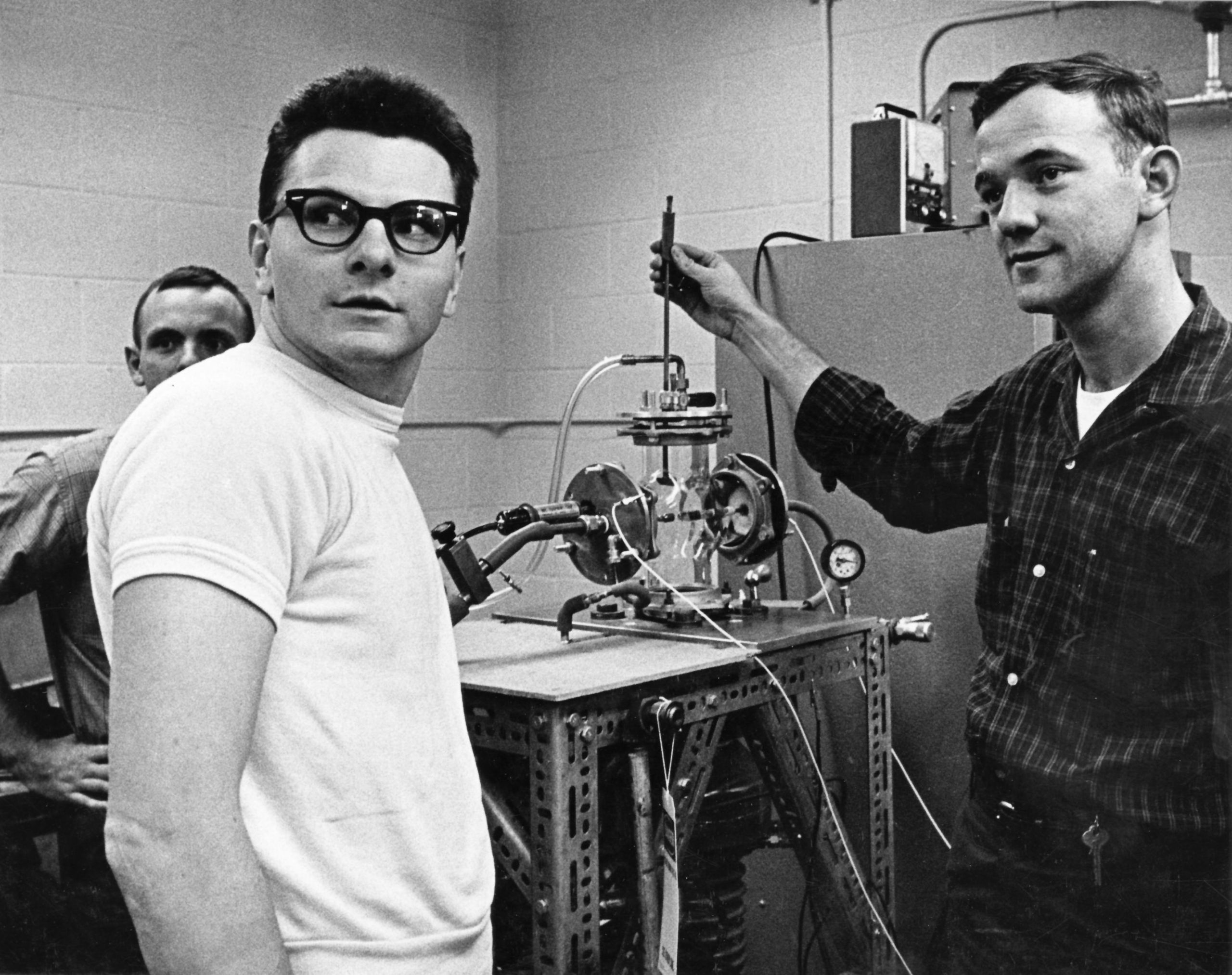 Black and white photo of three people in a lab. One holds a control to a complex metal device that sits in between them.