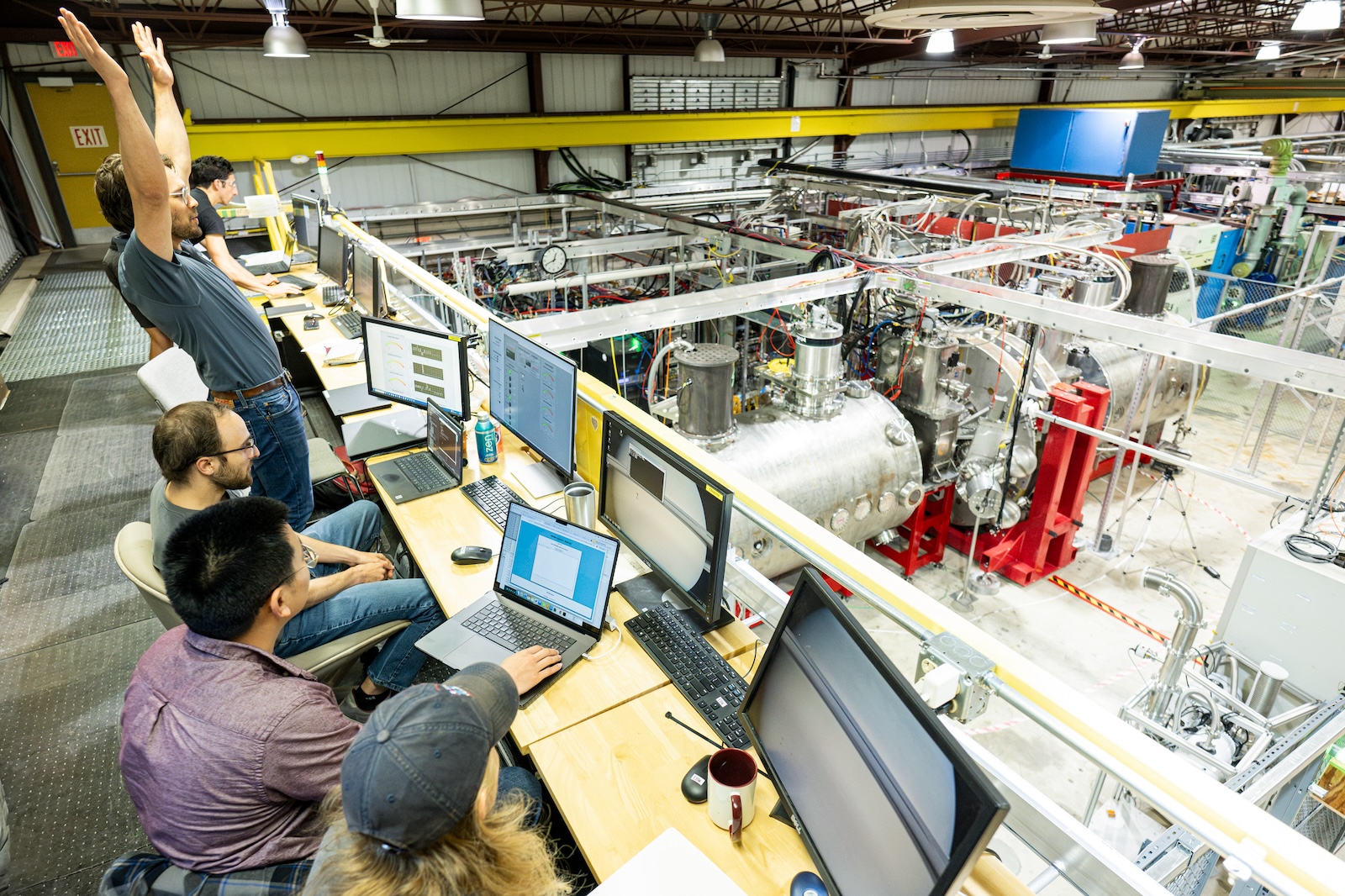 Five people sit around computer screens overlooking a large room filled with metal experimental equipment. One stands and raises their hands in celebration.