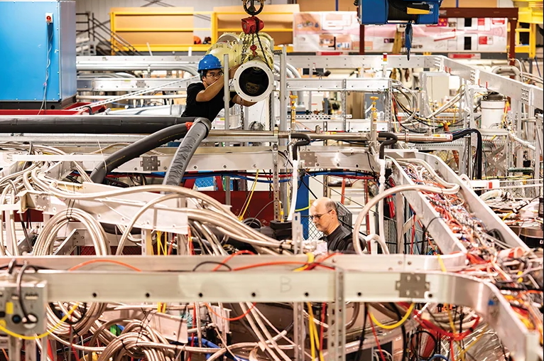 two people stand amongst large scientific equipment, including pipes and wires