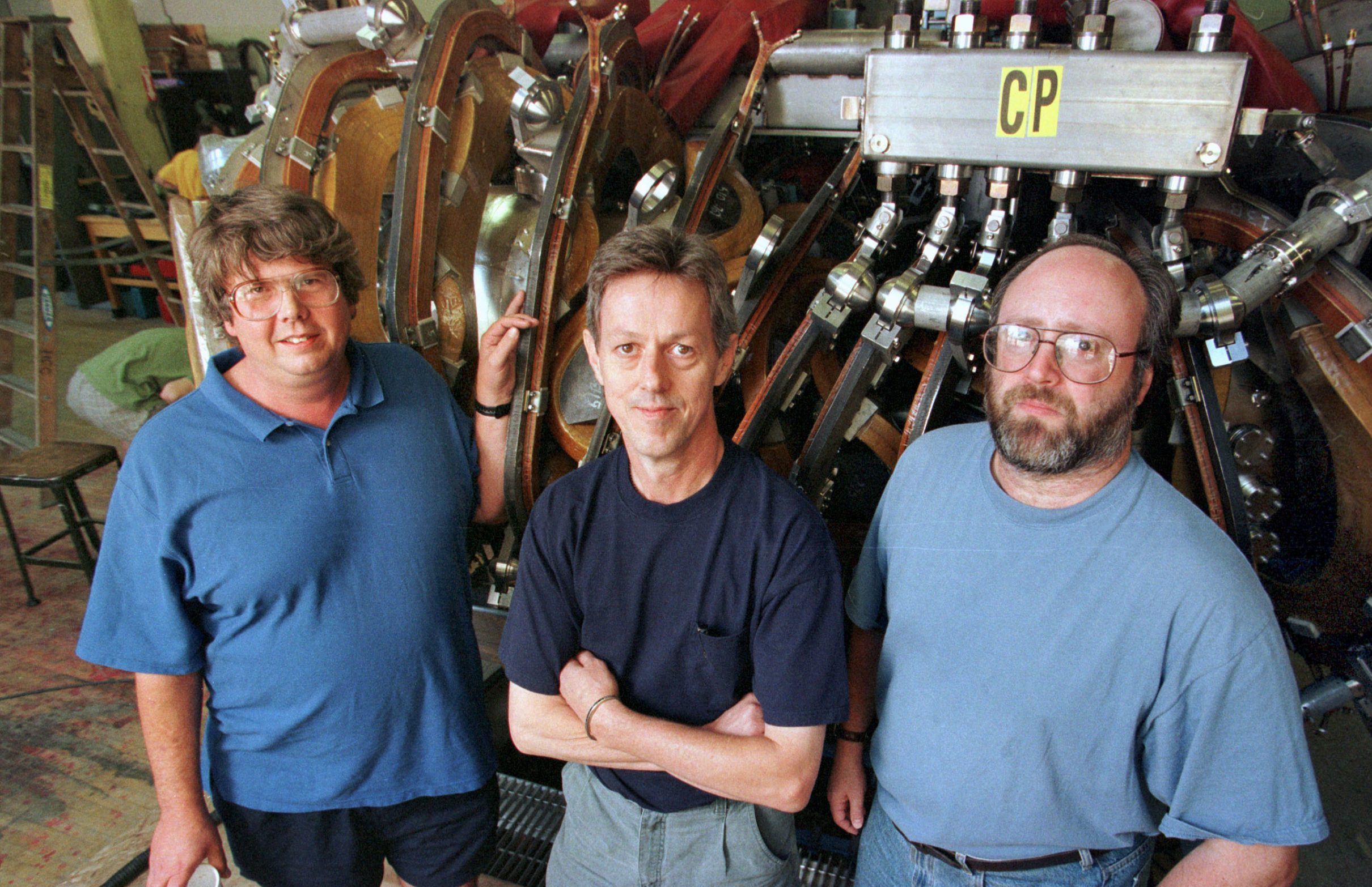 three white men wearing tshirts, the outer two wearing large glasses, standing in front of a large piece of scientific equpiment