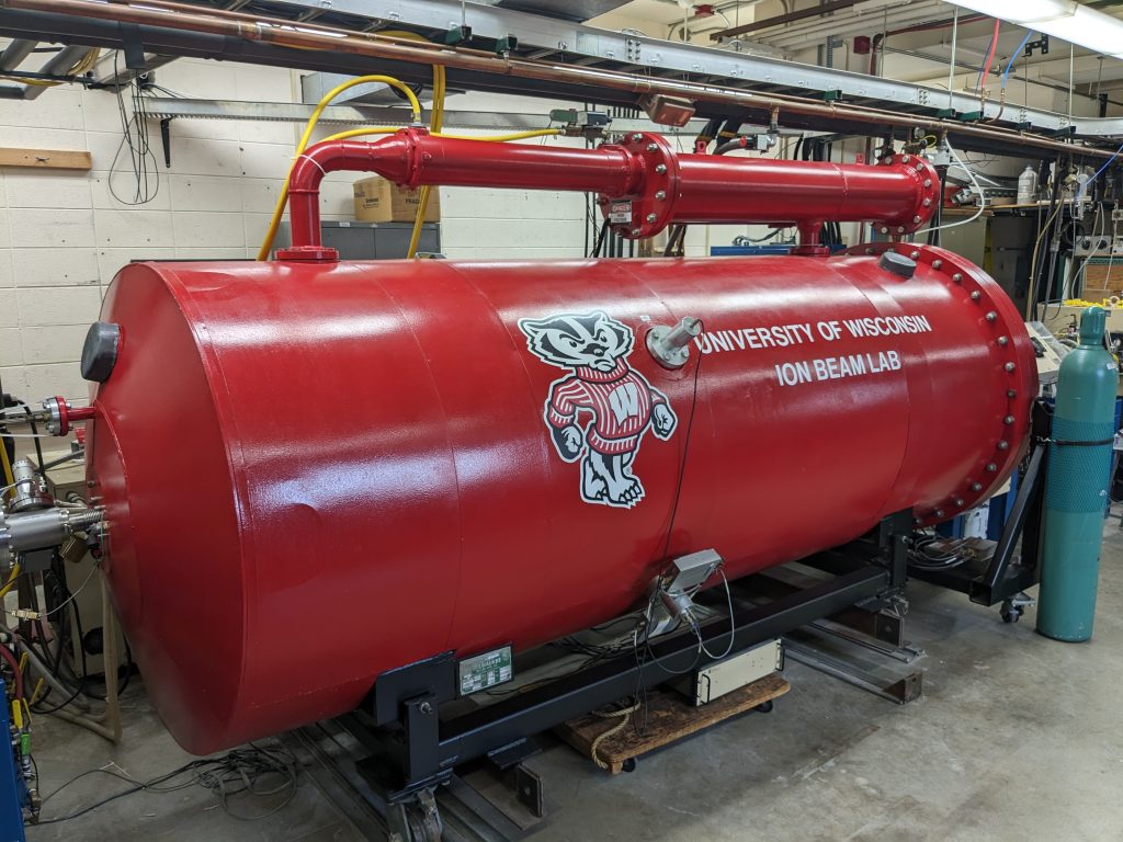 A large red cylinder with a red Bucky Badger paint job, in a large room with other pipes and scientific equipment.