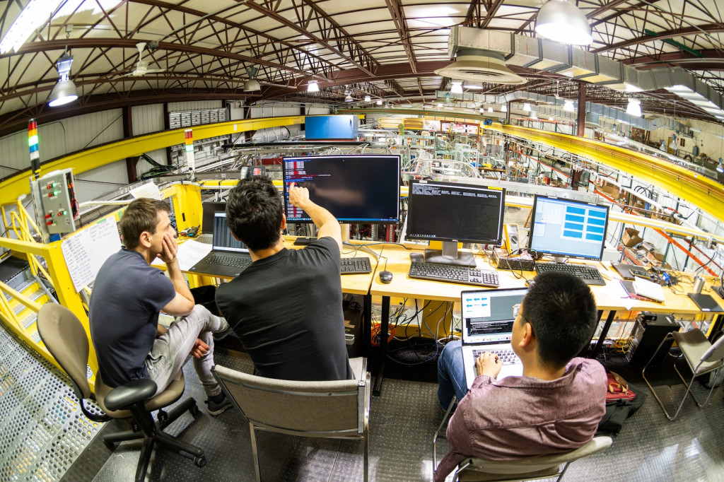 three men sit on an elevated platform with computer screens, overlooking a large room with lots of scientific equipment